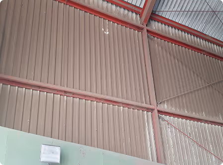 Innenwand einer Halle mit grauen Metallpaneelen und roten Stahlträgern, darüber ein Glasdach.